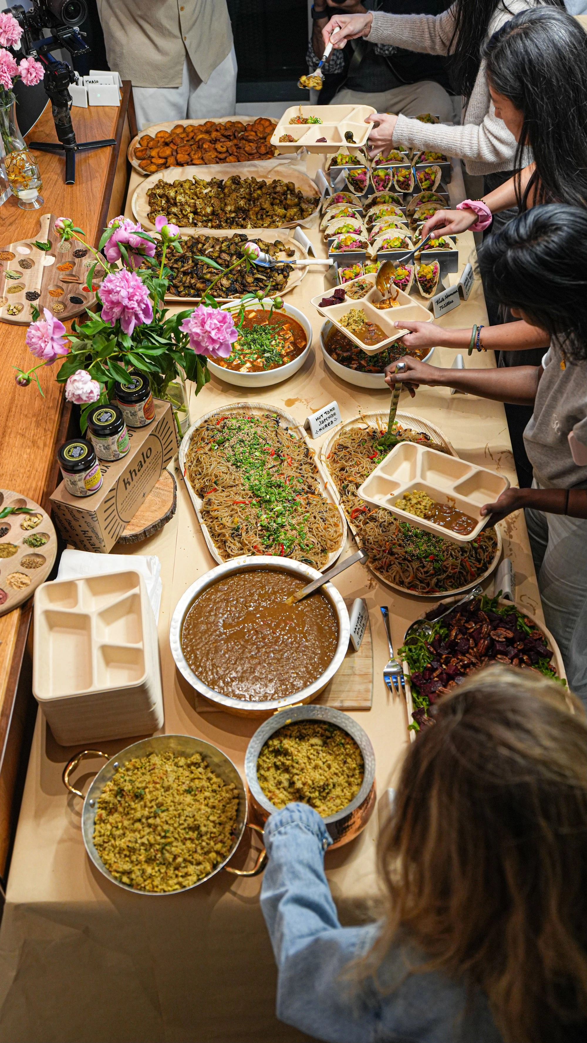 A long table of shared dishes, noodles, tacos, salads and peonies, with Khalo jars at the left edge