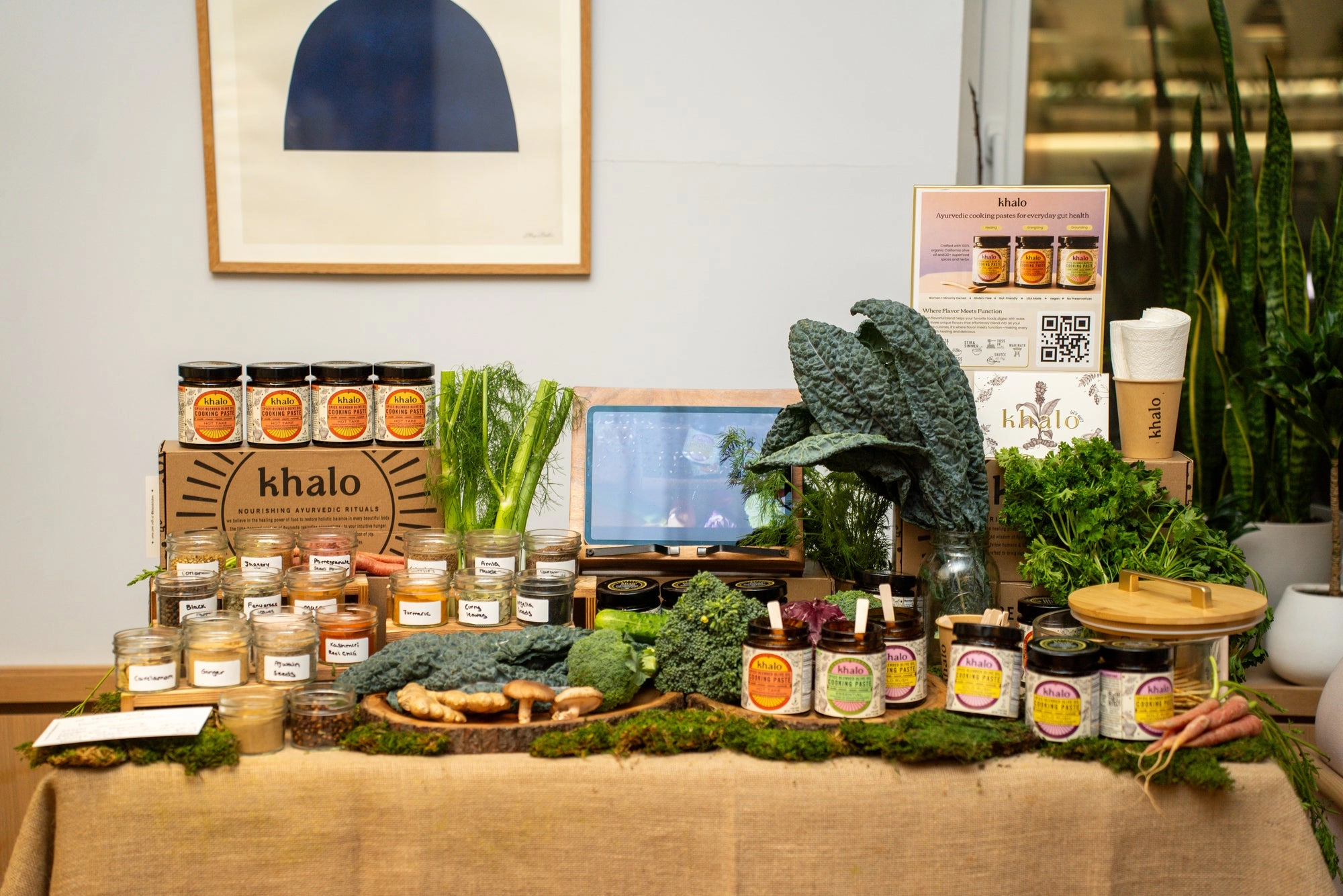 A Khalo display table with jars, spice bottles, kale, fennel and herbs on a burlap cloth