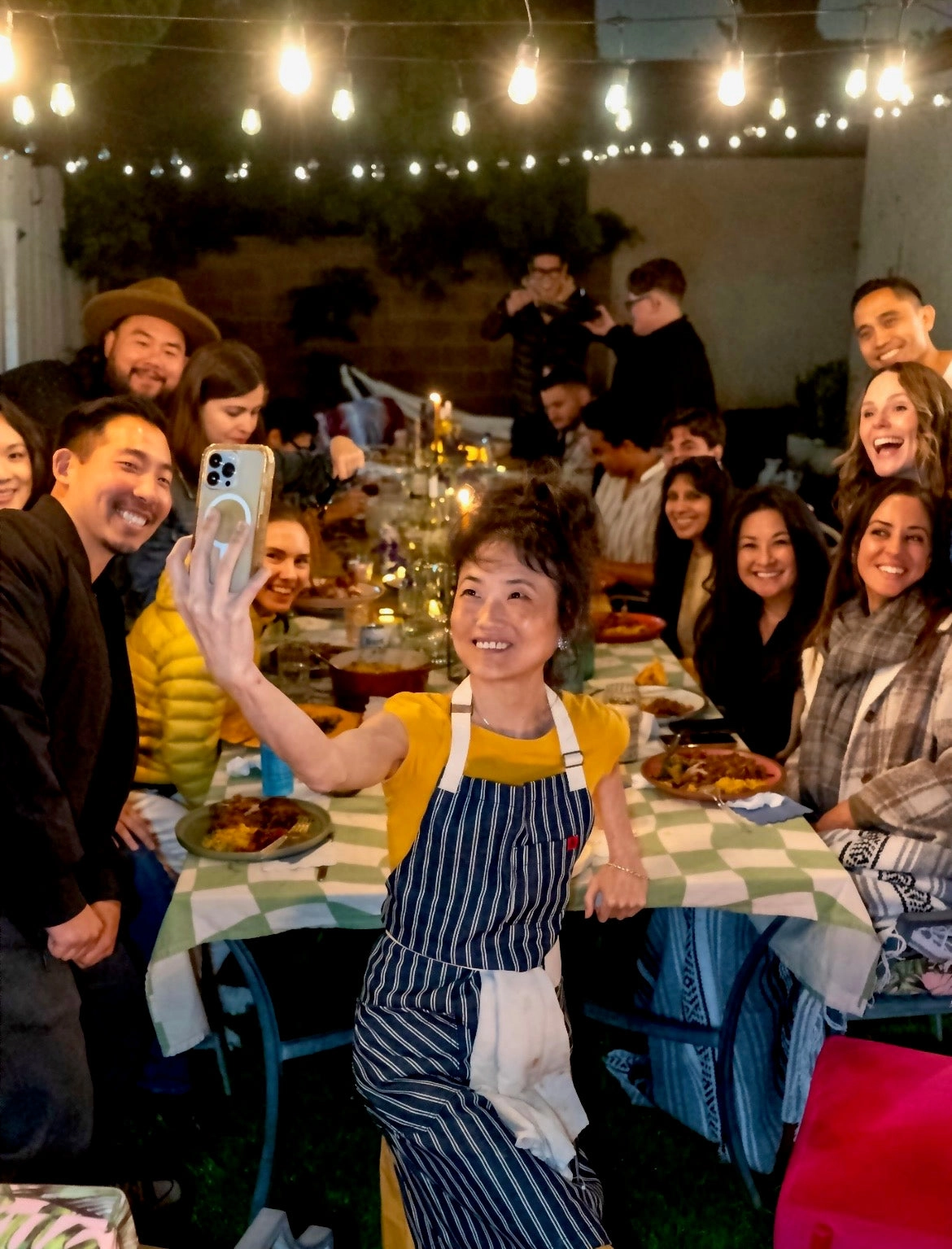 Friends laughing around a crowded dinner table, one person in an apron holding up a plate