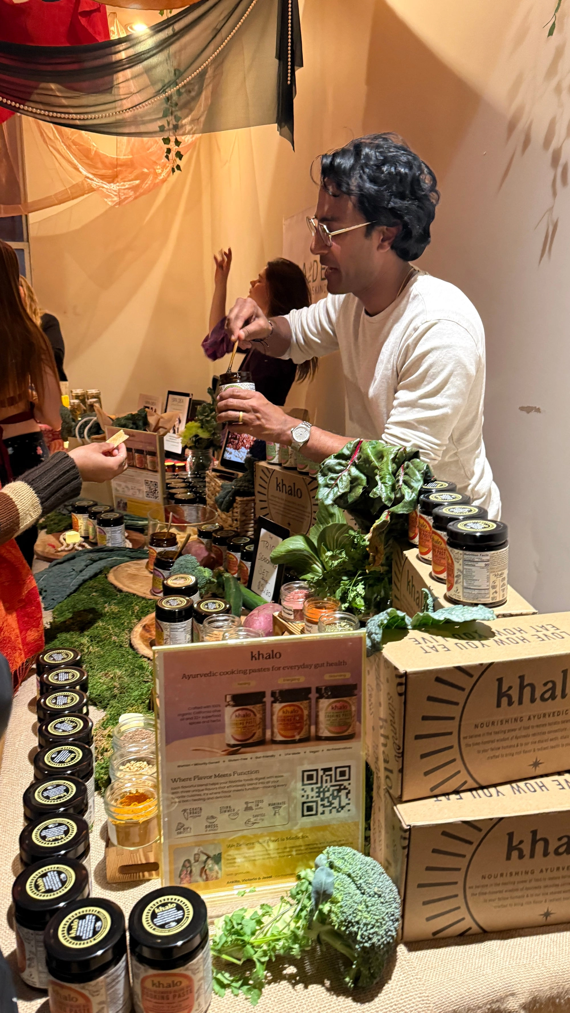 A cook at a market table lined with Khalo jars and greens